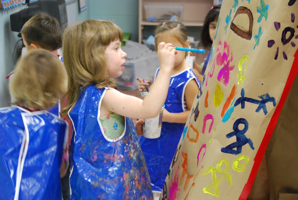 A prekindergartner wearing a blue smock paints at an easel